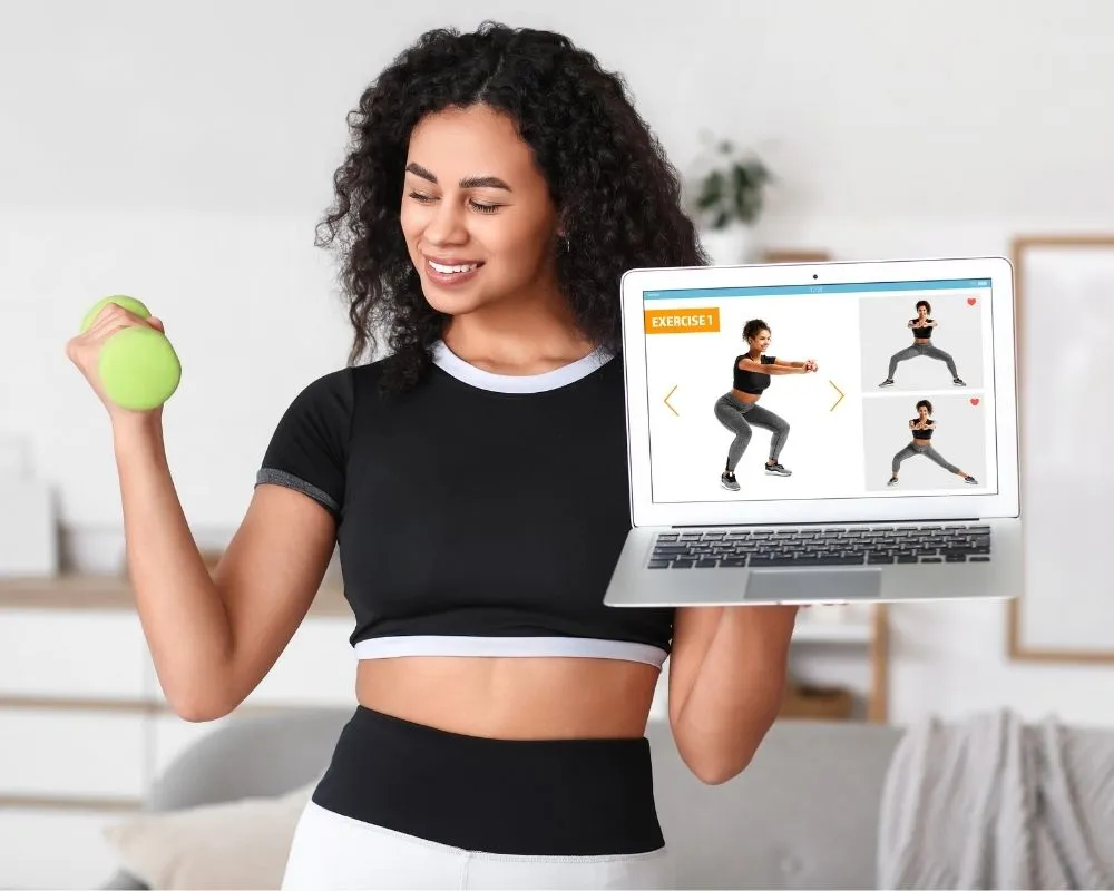 A woman following a home workout on a laptop while holding a dumbbell.
