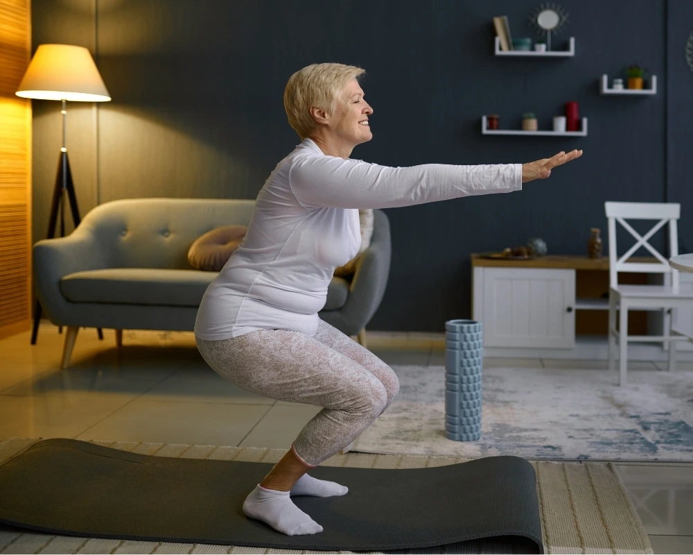 A smiling older woman doing a bodyweight squat on a mat in her living room.